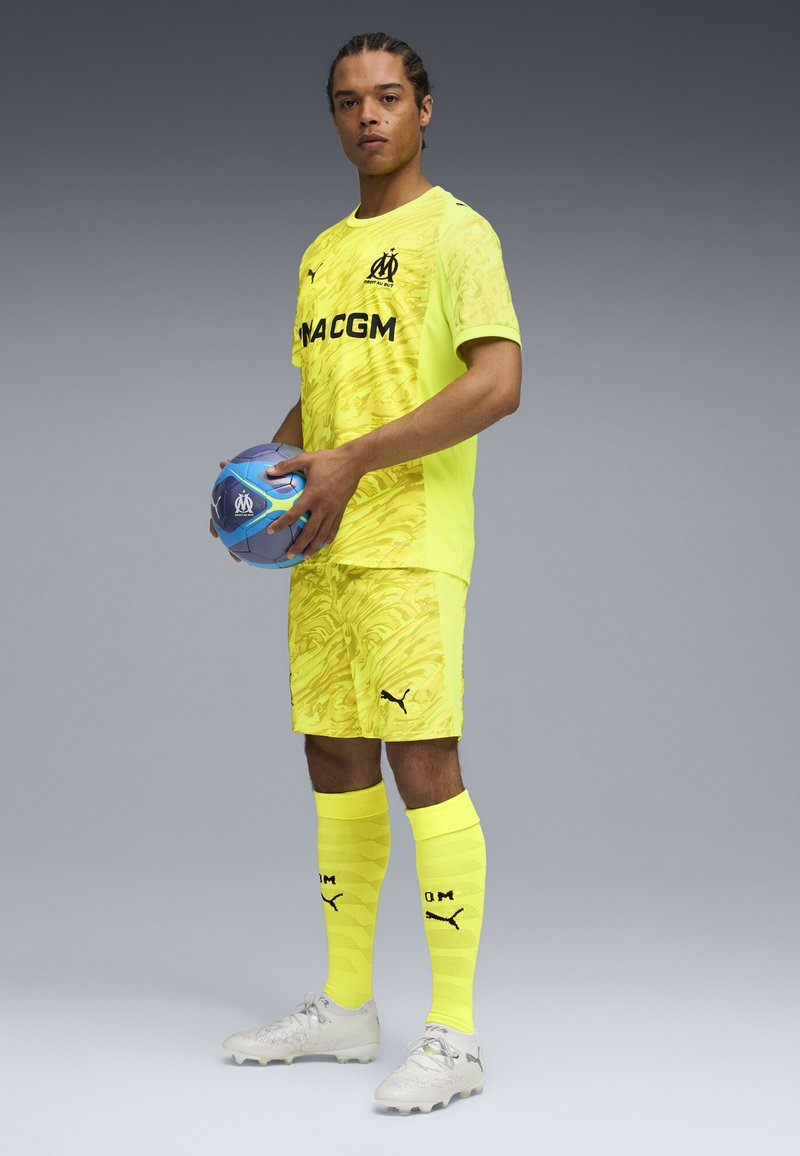 Uniforme da calcio giallo brillante con un motivo ondulato, abbinato a calze coordinate e scarpe da calcio grigie. Il giocatore tiene in mano un pallone da calcio blu.