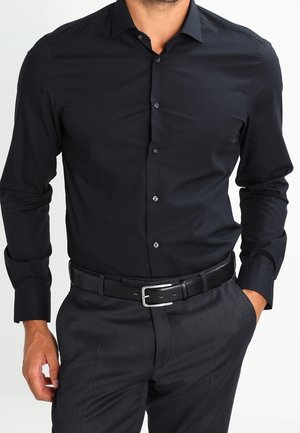 Man wearing a fitted black dress shirt tucked into dark gray trousers with a black leather belt, standing with one hand in pocket.