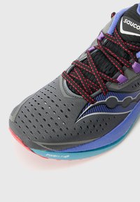 Chaussure de course en maille grise avec des accents noirs. Présente des lacets rouges, des détails violets et une semelle bleue avec un bout rouge et un motif.