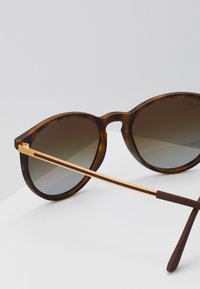 Óculos de sol tartaruga com lentes em gradiente castanho. Apresentam detalhes em metal dourado nas hastes e uma forma arredondada.
