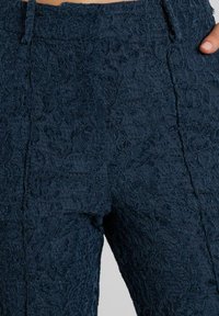 Pantalones de encaje azul marino con un patrón floral texturizado, cinturilla alta y diseño de pierna recta, que enfatizan el detallado intrincado y la artesanía.