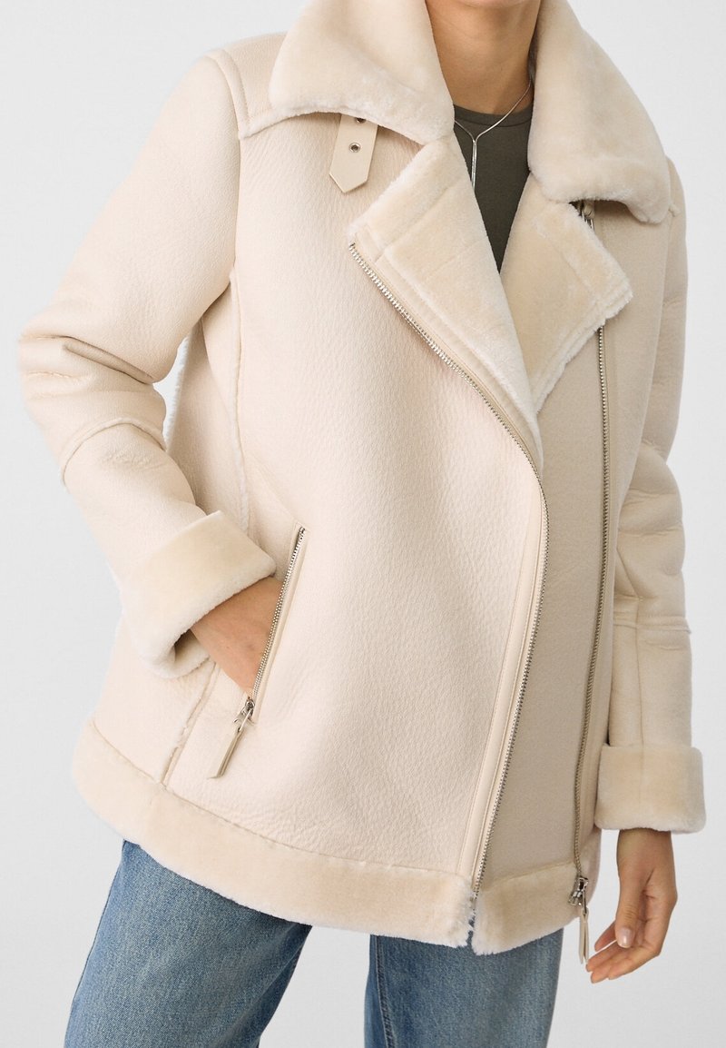 Giacca in shearling beige con colletto ampio, zip laterali e texture morbida. Presenta accenti lisci a contrasto lungo i bordi.