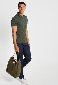 Olive green polo shirt, short sleeves, and a pocket. Navy trousers with a textured pattern. Carrying an olive tote bag with tan handles.