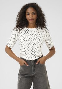 Haut à manches courtes blanc à pois, avec un col rond, associé à un pantalon en denim gris foncé taille haute. La texture semble lisse et décontractée.