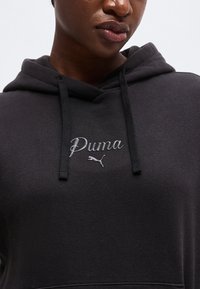 Personne portant un sweat à capuche noir avec le logo "Puma" et des cordons, montrant le haut de la poitrine et la partie inférieure du visage avec une expression neutre.