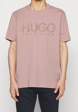 Man draagt een mauve Hugo Boss T-shirt met een geperst "HUGO HUGO BOSS" logo, gecombineerd met donkeren broek, getoond van nek tot heupen.