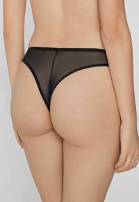Tanga negro de malla con una cinturilla elástica suave, que proporciona una cobertura mínima y presenta una textura semitransparente, delineada por bordes delgados.