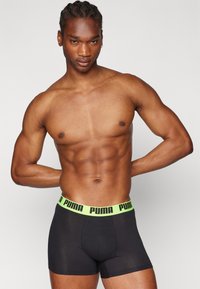 Fekete boxernadrág neon zöld derékpánttal, amelyen a Puma logó található. Az anyag simának tűnik és a testhez simul.
