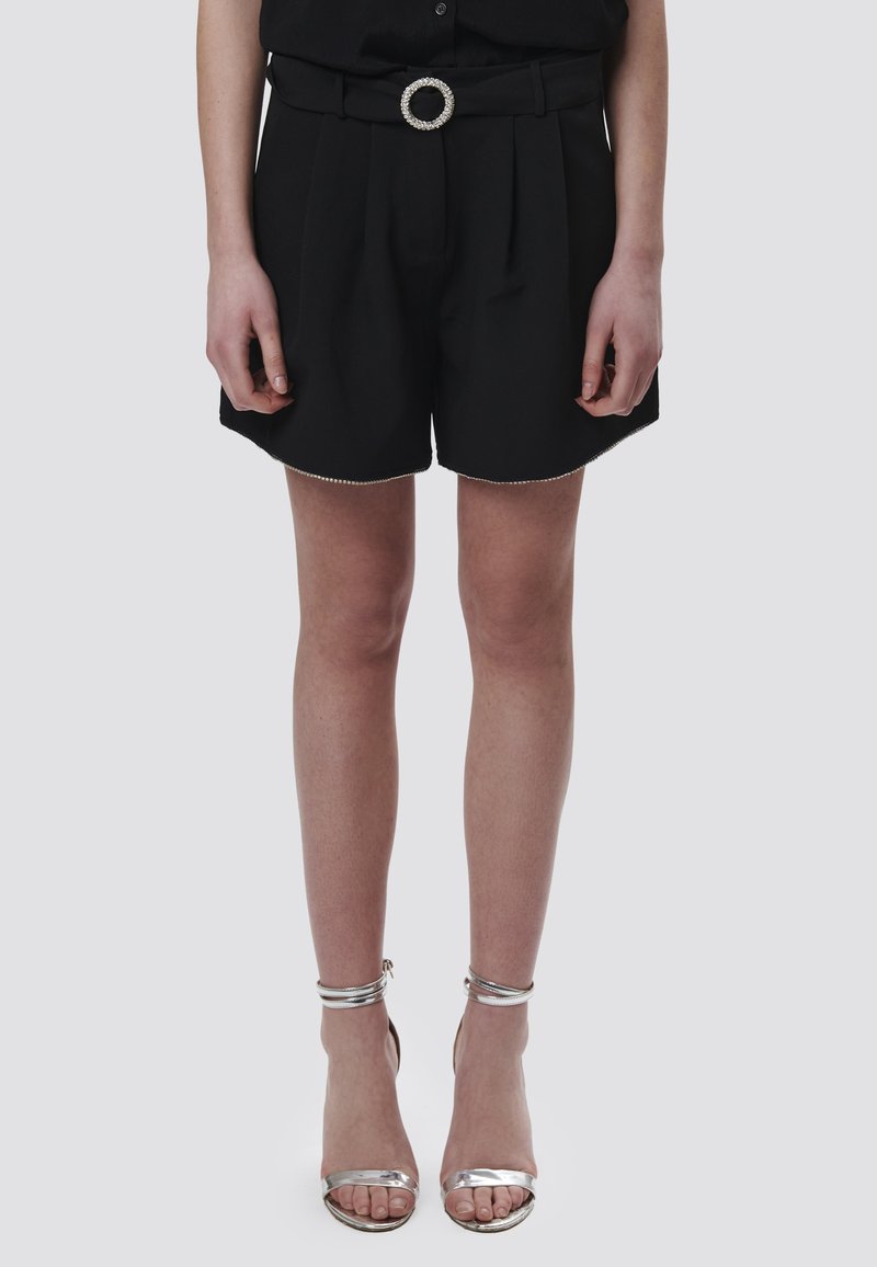 Pantalones cortos negros con pliegues, cinturilla ancha y hebilla de cinturón circular decorada con joyas. Combinados con sandalias plateadas de tacón alto y con tiras. Tejido suave.