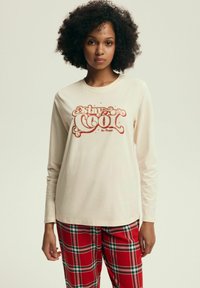 Beżowa bluzka z długim rękawem z czerwonym nadrukiem "stay COOL" i "Be Happy." Noszona z spodniami w kratę w czerwono-zielonym wzorze.