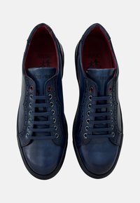 Las zapatillas de cuero azul oscuro presentan una textura suave, punta redonda y siete pares de ojales. El interior tiene un forro rojo y un detalle de marca.