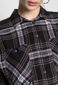 Camicia in flanella a quadri bianco e nero con bottoni, colletto classico e due tasche sul petto. Texture morbida con un motivo a quadri.