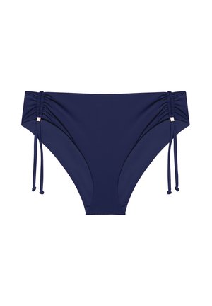 Donna con capelli ricci che indossa un bikini blu con dettagli dorati, in piedi davanti a uno sfondo chiaro uniforme, sorridente verso la macchina fotografica.