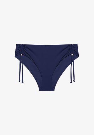 Femeie cu păr creț care poartă un costum de baie albastru cu accente aurii, stând în fața unui fundal simplu și luminos, zâmbind către cameră.