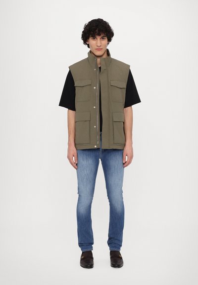 Olijfgroene mouwloze vest met een hoge kraag, voorzakken en een rits, gecombineerd met een zwart t-shirt en lichtblauwe jeans.