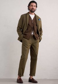Oliwkowa marynarka na brązowej kamizelce, biała koszulka i oliwkowe spodnie. Teksturowane ciemno-brązowe buty. Luźny fason, podwinięte nogawki spodni.