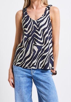 Vrouw draagt een mouwloos, zwart-wit zebraprint top met gerimpelde bandjes en lichtblauwe spijkerbroek, tegen een effen achtergrond.
