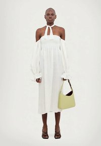 Γυναίκα με λευκό μίντι φόρεμα χωρίς ώμους, με μακριά φουσκωτά μανίκια, κρατώντας μια ανοιχτόχρωμη πράσινη τσάντα και φορώντας μαύρες λουρίδες ψηλές γόβες.