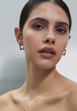 Kvinde med glat hud og brune øjne iført gyldne geometriske øreringe og en lille ørestik, med håret trukket tilbage mod en neutral baggrund.
