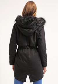 Kobieta z długimi włosami, ubrana w czarną kurtkę parka z kapturem i sztucznym futrem, stojąca tyłem na jednolitym tle.