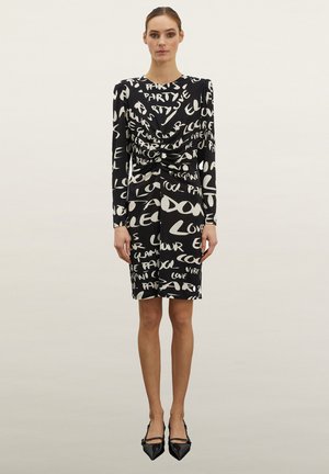 Vestido negro con diseño de texto manuscrito en blanco, de mangas largas, con fruncido en el frente, falda a la altura de la rodilla, combinado con zapatos negros de puntera puntiaguda.