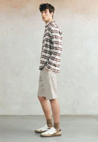 Młody mężczyzna stoi z rękami w kieszeniach, ubrany w brązową koszulę w kratę, beżowe szorty, brązowe skarpetki i białe trampki, na tle gładkiej ściany.