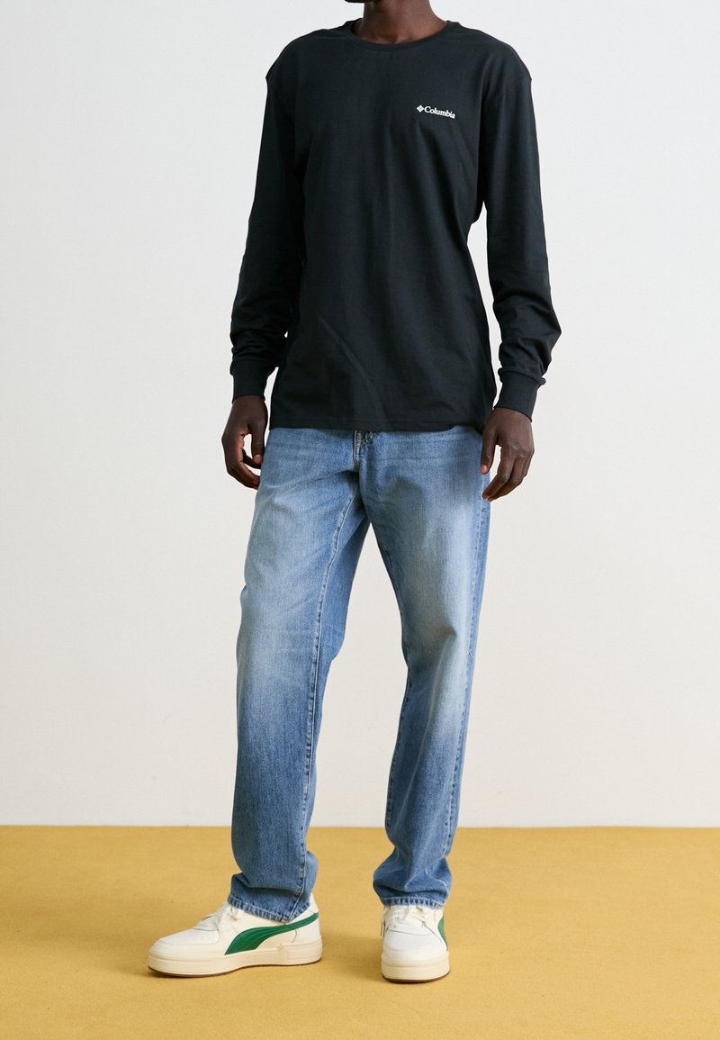 Personne portant un t-shirt Columbia noir à manches longues, un jean bleu clair et des baskets blanches avec des rayures vertes sur un sol jaune.