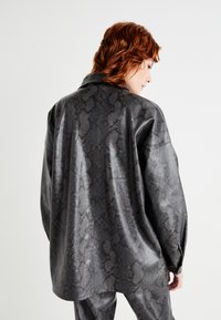 Schwarze Kunstlederhemden mit strukturiertem Schlangenprint, lockerer Passform, spitzen Kragen und langen Ärmeln. Die Rückansicht zeigt eine minimale Tailoring.