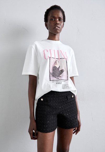 T-shirt blanc à manches courtes avec un graphisme "CELINE" en rose et une impression photo; associé à un short noir texturé avec des boutons dorés.