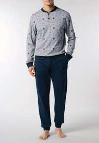 Uomo che indossa una maglietta henley a maniche lunghe grigia con piccoli motivi di uccelli e pantaloni jogger blu navy, in piedi a piedi nudi su uno sfondo bianco.