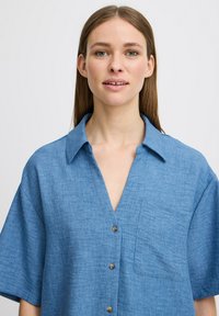 Camicia di lino blu con vestibilità rilassata, maniche corte, scollo a V e chiusura con bottoni. Presenta una tasca sul petto e un tessuto strutturato.