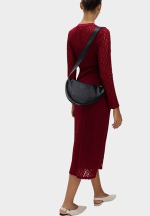 Robe en dentelle rouge tricotée avec des manches longues, longueur mi-mollet, accompagnée d'un sac à bandoulière en cuir noir en forme de demi-lune et de chaussures blanches à enfiler.