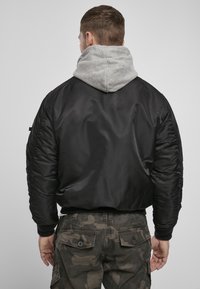 Jachetă bomber neagră cu mâneci capitonate, glugă gri în contrast, manșete ribbate și o textură netedă și lucioasă. Pur tà pe un pantalon cargo cu imprimeu de camuflaj.