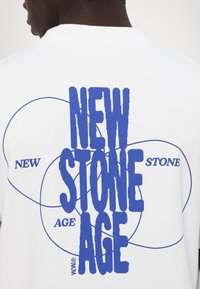 Hvit bomull T-skjorte med fet blå tekst som sier "NY STEINALDER." Sirkulære mønstre i blå omgiver teksten. Enkel og grafisk design.