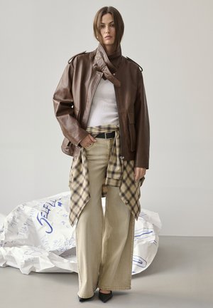 Frau trägt eine braune Lederjacke über einem weißen Oberteil, mit einem karierten Hemd, das um weite beige Hosen gebunden ist, und schwarzen spitzen Schuhen.