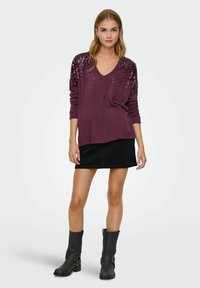 Weinroter Pullover mit V-Ausschnitt, verziert mit Pailletten an den Schultern, kombiniert mit einem schwarzen Rock und kniehohen schwarzen Stiefeln.