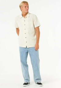 Kortærmet beige skjorte med knapper og lomme, parret med lyseblå jeans og sorte sko med hvide såler. Glat stoftekstur.