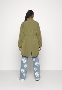 Persona con trenzas, vistiendo una chaqueta larga acolchada de color verde oliva y pantalones vaqueros azules con estampado de flores blancas, de pie, de espaldas.