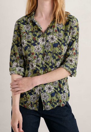 Woman wearing green button-up shirt with white floral pattern and rolled-up sleeves, paired with dark blue jeans, standing with arms crossed.