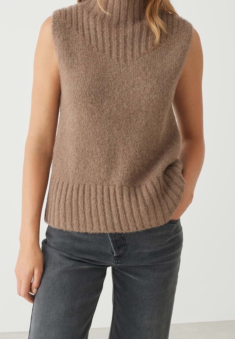Beiger ärmelloser Rollkragenpullover aus weichem, strukturiertem Stoff mit geripptem Kragen und Saum, kombiniert mit dunkelgrauen Jeans.