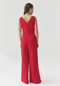 Rode jumpsuit met brede pijpen, mouwloos ontwerp en een diepe V-rug. Gemaakt van textuurstof met een getailleerde taille en naadloze details.