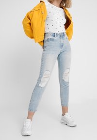 Ljusblå rivna jeans med hög midja, i kombination med en vit skjorta med prickar och en ljusgul jacka, kompletterat med vita sneakers.