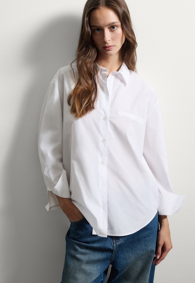 Camisa blanca de botones con cuello, bolsillo en el pecho y mangas largas, combinada con pantalones vaqueros de mezclilla azul claro. Tejido de algodón texturizado.