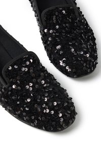 Zapatos negros sin cordones cubiertos de brillantes lentejuelas negras. Cuentan con un upper de tela suave y un diseño de punta redondeada con detalles elásticos.