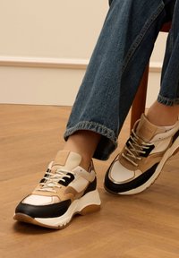 Sneaker con un mix di materiali beige, neri e bianchi; suola spessa; trame in mesh, pelle e camoscio; lacci e marchi in evidenza.