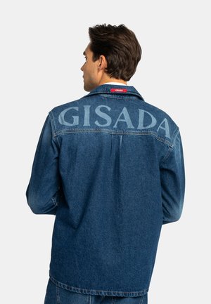 Mann trägt eine dunkelblaue Denimjacke mit großem verblasstem "GISADA" Text auf dem Rücken und einem kleinen roten Etikett am Kragen, er ist abgewandt.