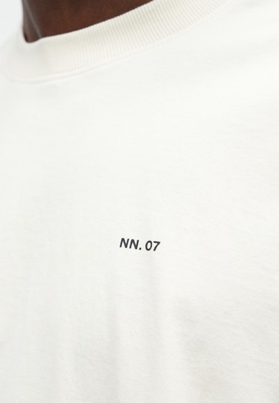 Lähikuva valkoisesta t-paidasta, jossa on pieni musta "NN.07" -teksti painettuna rinnassa kaulan lähellä.