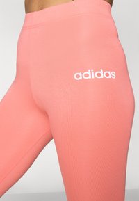 Roza legging iz raztegljivega materiala, z visokim pasom in belim logotipom "adidas" na levi stegni. Gladka tekstura in prilegajoč dizajn.