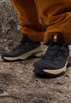Chaussures de trail noires et beiges portées avec un pantalon marron, debout sur un sol rocheux en plein air.