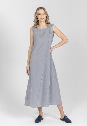 Kobieta stojąca z jedną ręką na biodrze, ubrana w bezrękawnikową, paskowaną sukienkę midi oraz granatowe wsuwane buty na jednolitym tle.